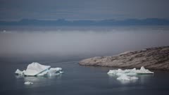 Icebergs move in icy water, fog, cloud layer hovers just above water, mountains in the distance