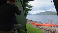 Man Setting Up The Interior Of A Tent Near Caledonian Canal In Scotland With Orange Kayak On The Shore - Medium Shot