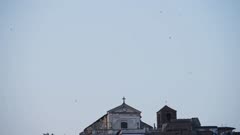 Flock Of Birds Flying Over The Historical Santa Maria Assunta Church In The Small Village In Anguillara On Bracciano Lake, Lazio, Rome, Italy. - wide shot