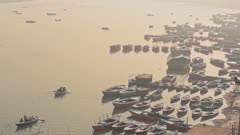 Boats Docked At The Near The Riverbank In Varanasi, India With Two Boats Sailing Over The River Ganges On A Sunrise - Aerial Shot