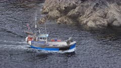 Medium shot of small fishing trawler traversing through frame as it enters a rocky cove. Cornwall, UK.