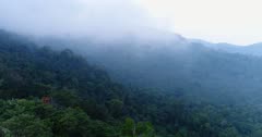 Drone footage of the Bokor National Park near Kampot, Cambodia. The camera is going sideways showing the untouched forest covering the hills of the Bokor National Park. A thick fog is covering the trees and moving fast over them.