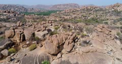 Drone footage of the UNESCO World Heritage temple complex area in Hampi, Karnataka, India, with the rocky landscape and bright green rice fields on the other side of the Tungabhadra river, hills in the background. The camera is going backward while descending over the rocky landscape.