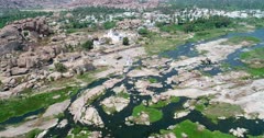 Drone footage of the countryside near Hampi, Karnataka, India, with the rocky bed of the Tungabhadra river, the granite like boulders forming hills along it, the Chinthamani Temple and the Anegundi village behind. The camera is going sideway over the river turning around the temple.