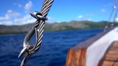 Footage of the end of steel cable making a loop and hook tighting it, a tropical island with rocky coast and palm trees is in the background, Forgotten Islands, Indonesia. The camera is staying still focusing first on the cable and then on the background.