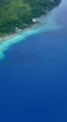 Vertical drone footage of a diving boat floating in front of a deserted beach surrounded by tropical vegetation and colorful shallow reef in the north of Dawera island, Indonesia. The camera is facing the north-west coast of the island and is panning towards the north-east.