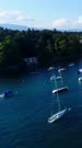 Vertical drone footage of the shore of the Leman lake in Genthod with a lot of small sailing boats. The camera is going over the water along the shore following the boats and slowly panning towards them while tilting down.