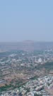 Vertical drone footage of the Marble Mughal mausoleum Bibi Ka Maqbara in Aurangabad, Maharashtra, India, with the city low buildings mostly white around and some hills in the background. The camera is going over the city towards the monument.