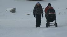People With Baby Stroller Walk Through Village In Rugged Snowy Area At Base Of Mountain