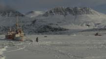 People Walk Next To Boats Stuck In Ice At Base Of Mountains