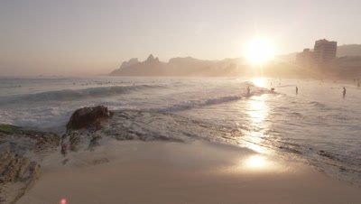 Slow motion, sunset shot of white waves rolling onto beach as people swim
