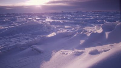 Scenic - Slow pan across foreground snow that has been shaped and sculpted by winds.  Focus on foreground, pan stops to reveal intricate snow shapes.  Rugged ice stretching to the horizon in background, where an out-of-focus sun sets behind a cloud bank.  Low Angle.  Wide.