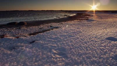 Scenic  - Pan across polar bear tracks in faceted snow along shore line as sun rises on the horizon.  Ext. Wide.