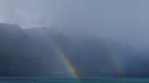 Double Rainbow Over Lake In Cloudy, Windy Conditions With Mountain Backdrop.