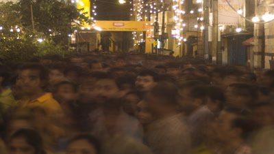 Medium wide angle over heads of densely packed people as they stream through a floodlit street during the festival of Durga Puja