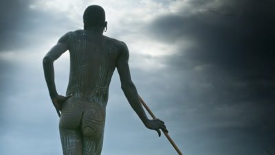 Close up Suri tegay, young man, stick fighter, back view as stands still leaning on his fighting stick looking into distance with dramatic storm clouds growing gradually darker