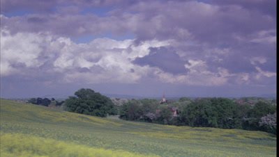 Medium wide angle looking across yellow rapeseed field in full flower to English woodland, distant hills and church spire with building clouds overhead