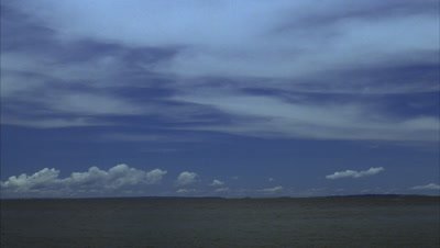 Wide angle blue sky with small fluffy white clouds and large expanses of wispy cloud streaming over grey water clearing to blue sky